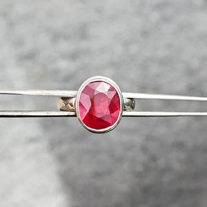 May include: A silver ring with a large, oval-cut ruby gemstone. The ruby is a bright red hue, set in a simple, polished silver band. The ring is held by tweezers against a blurred background.