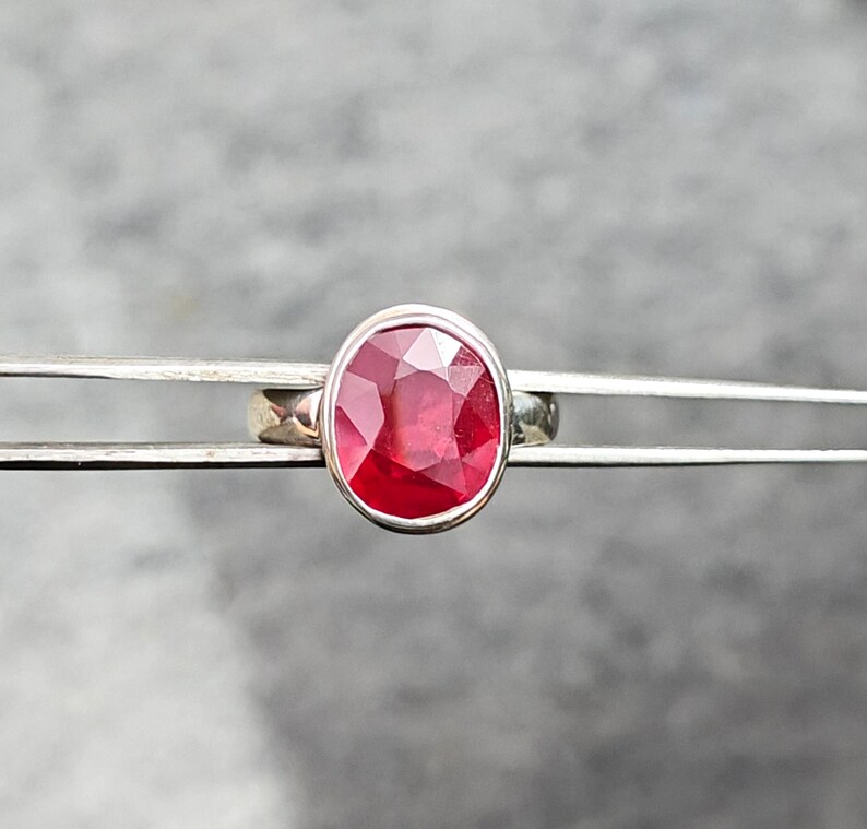 May include: A silver ring with a large, oval-cut ruby gemstone. The ruby is a bright red hue, set in a simple, polished silver band. The ring is held by tweezers against a blurred background.