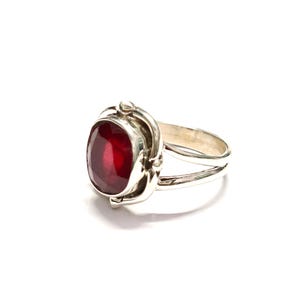 Puede incluir: Anillo de plata con una gran gema roja oscura de corte ovalado. La gema est&aacute; engastada en un engaste de plata decorativo con detalles ornamentados. El anillo tiene un dise&ntilde;o de ca&ntilde;a dividida. Sobre fondo blanco.