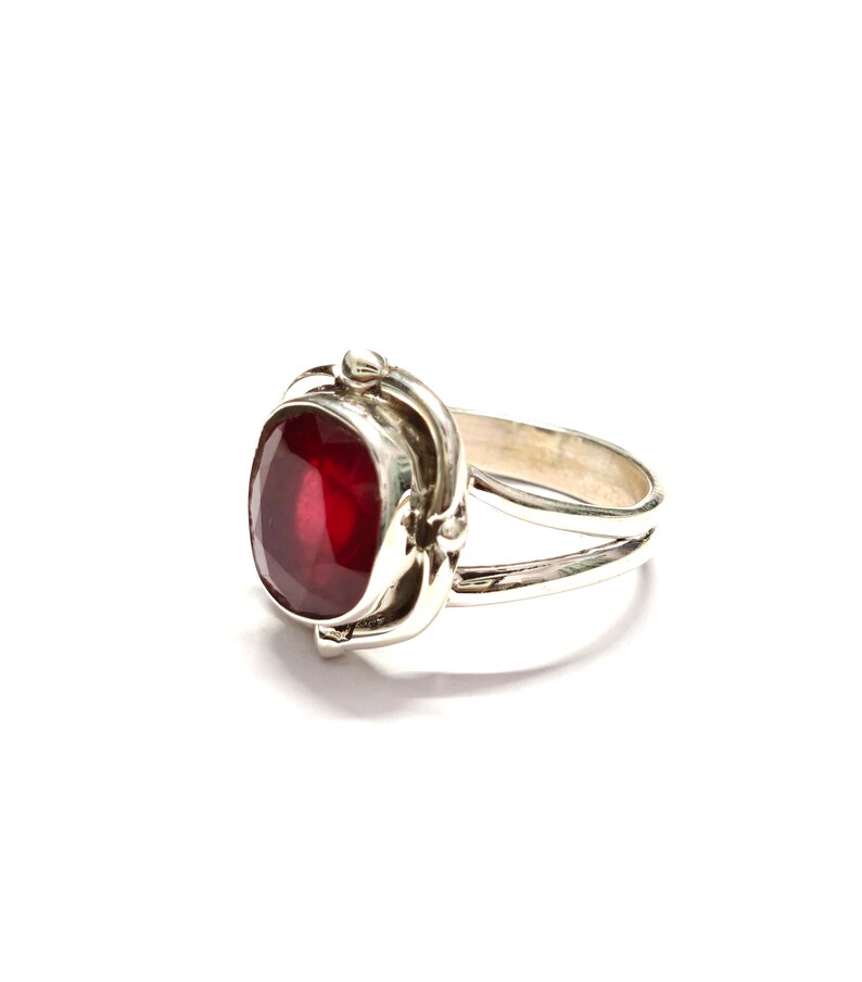 Puede incluir: Anillo de plata con una gran gema roja oscura de corte ovalado. La gema est&aacute; engastada en un engaste de plata decorativo con detalles ornamentados. El anillo tiene un dise&ntilde;o de ca&ntilde;a dividida. Sobre fondo blanco.