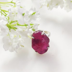 May include: A silver ring featuring a large, deep pink, irregularly shaped gemstone. The ring is displayed against a white background with white flowers.