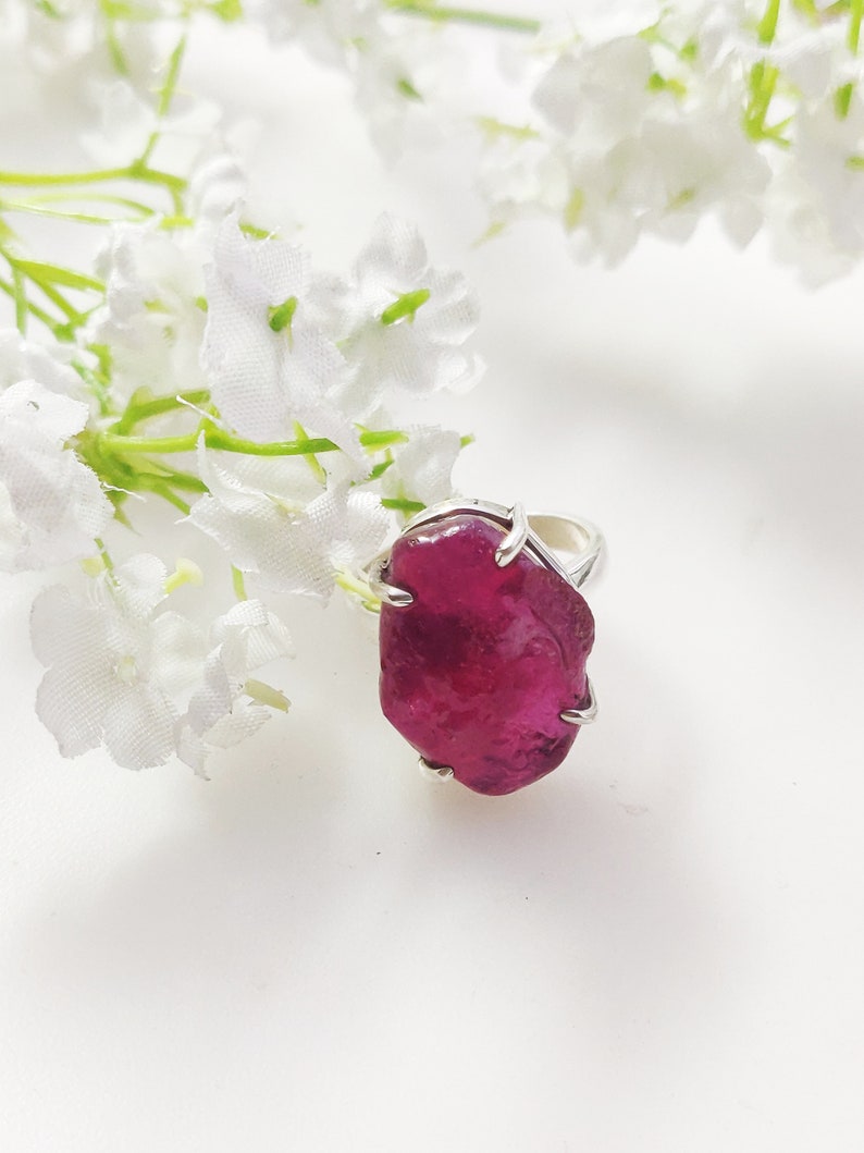 May include: A silver ring featuring a large, deep pink, irregularly shaped gemstone. The ring is displayed against a white background with white flowers.