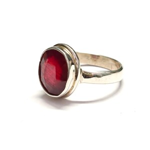 May include: A silver ring with a large, oval-cut, deep red gemstone. The gemstone is set in a bezel setting with a double band design. The ring is shown against a white backdrop, accentuating the polished metal and the vibrant colour of the stone.