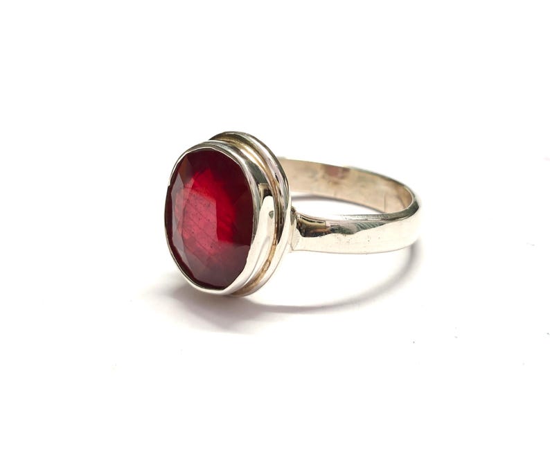 May include: A silver ring with a large, oval-cut, deep red gemstone. The gemstone is set in a bezel setting with a double band design. The ring is shown against a white backdrop, accentuating the polished metal and the vibrant colour of the stone.