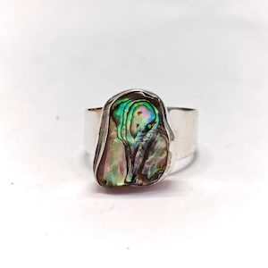 Puede incluir: Un anillo de plata con una gran concha de abulón iridiscente engastada en el centro. La concha tiene un patrón de color verde, azul y morado.