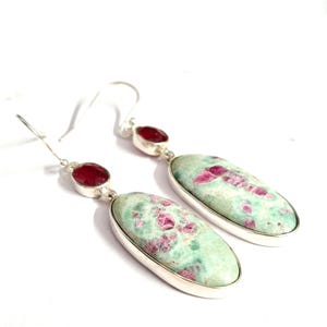 Pu&ograve; includere: Un paio di orecchini pendenti con pietre preziose ovali, verde chiaro con inclusioni rosa, incastonate in metallo argentato. Ogni orecchino ha una piccola gemma rossa in cima.