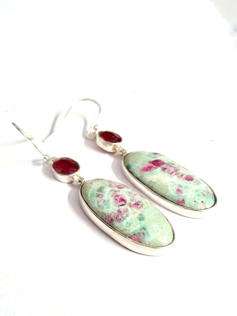 Pu&ograve; includere: Un paio di orecchini pendenti con pietre preziose ovali, verde chiaro con inclusioni rosa, incastonate in metallo argentato. Ogni orecchino ha una piccola gemma rossa in cima.