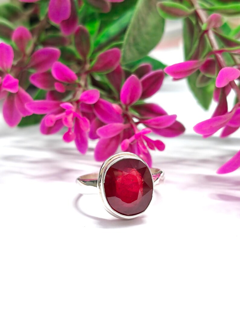May include: A silver ring featuring a large, oval-cut, deep red gemstone. The ring is set against a backdrop of pink flowers and green leaves. The gemstone is the focal point, showcasing its rich colour and reflective qualities.