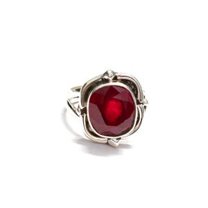 Puede incluir: Un anillo de plata con una gran piedra preciosa roja oscura de talla cuadrada. La piedra preciosa est&aacute; engastada en un bisel de plata decorativo con un dise&ntilde;o detallado. El anillo se muestra sobre un fondo blanco.