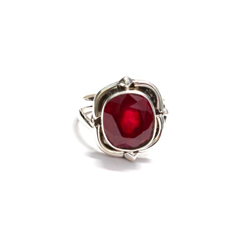 Puede incluir: Un anillo de plata con una gran piedra preciosa roja oscura de talla cuadrada. La piedra preciosa est&aacute; engastada en un bisel de plata decorativo con un dise&ntilde;o detallado. El anillo se muestra sobre un fondo blanco.