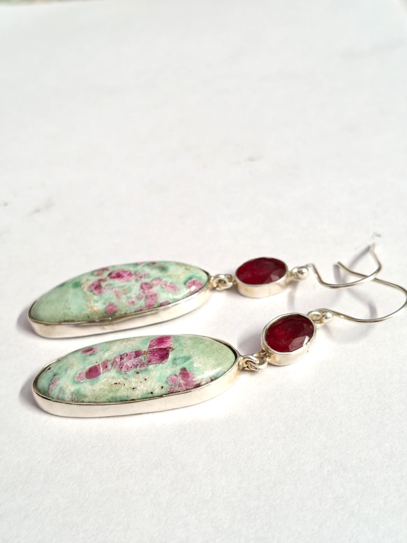 Pu&ograve; includere: Un paio di orecchini in argento. Ogni orecchino presenta una grande pietra ovale verde chiaro con inclusioni rosa, abbinata a una pietra rosso scuro pi&ugrave; piccola, di forma ovale. Gli orecchini sono incastonati in argento.