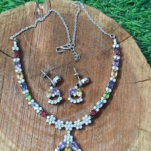 May include: A silver necklace and earring set with a variety of colourful gemstones. The necklace has a floral design with a pendant, and the earrings have matching gemstone clusters. The jewellery is displayed on a wooden surface.