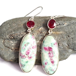 Puede incluir: Un par de pendientes de plata con piedras ovaladas de color verde claro con inclusiones rosas, y una pequeña piedra roja redonda. Los pendientes se exhiben sobre una roca gris.