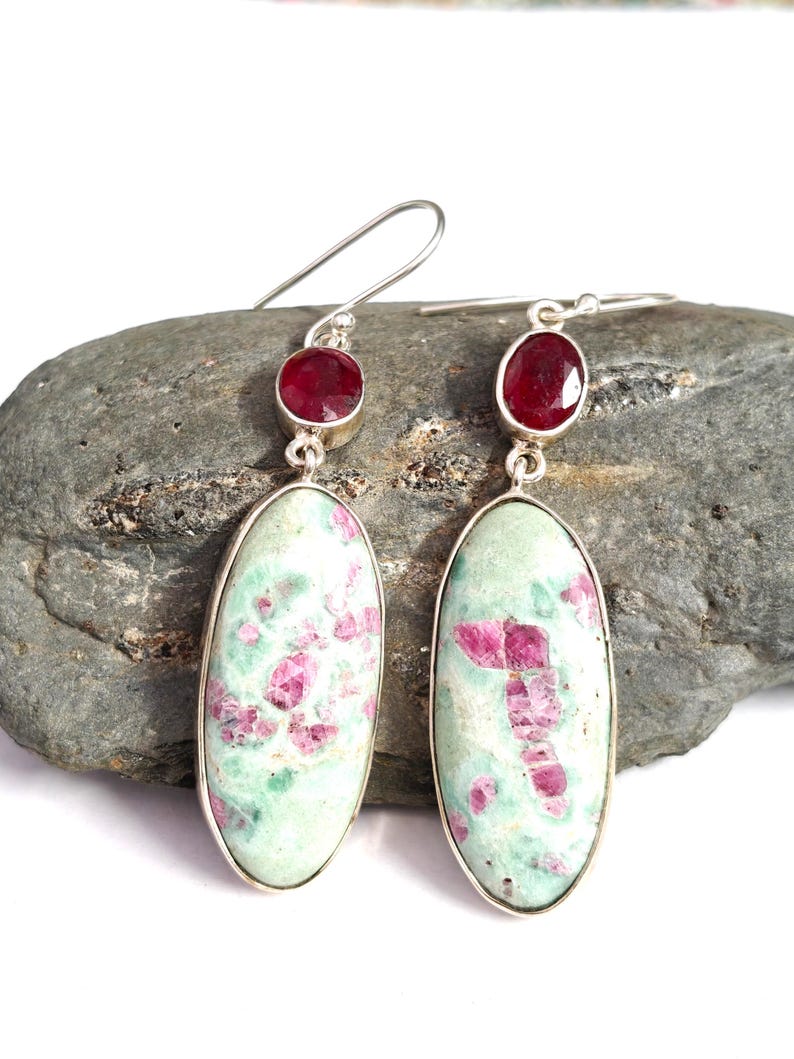 Pu&ograve; includere: Un paio di orecchini in argento con pietre ovali verde chiaro con inclusioni rosa e una piccola pietra rossa rotonda. Gli orecchini sono esposti su una roccia grigia.