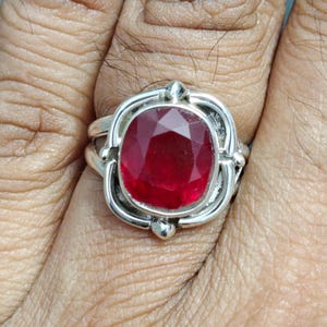 Puede incluir: Un anillo de plata con una gran piedra preciosa de rub&iacute; de corte ovalado. El rub&iacute; es de color rojo intenso y est&aacute; engastado en una montura de plata decorativa con detalles ornamentados. El anillo se muestra en un dedo.
