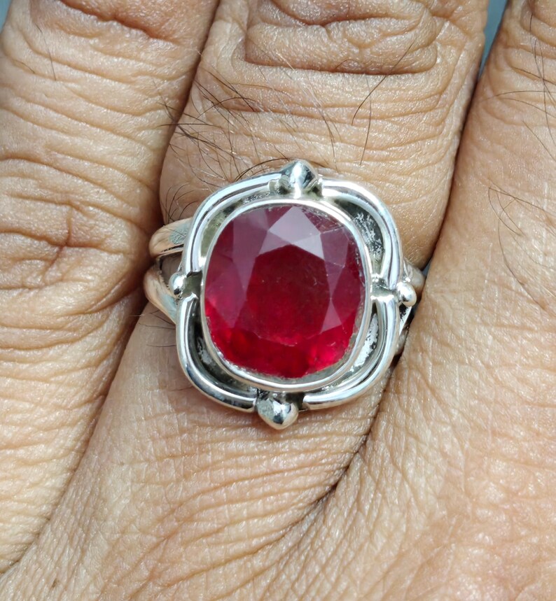 Puede incluir: Un anillo de plata con una gran piedra preciosa de rub&iacute; de corte ovalado. El rub&iacute; es de color rojo intenso y est&aacute; engastado en una montura de plata decorativa con detalles ornamentados. El anillo se muestra en un dedo.