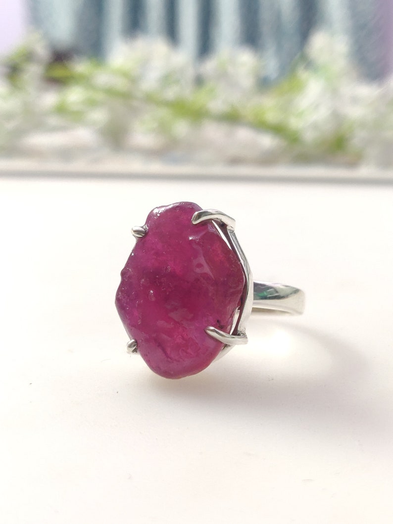 May include: A silver ring featuring a large, raw, pink ruby gemstone. The ruby is set in a simple, prong setting.