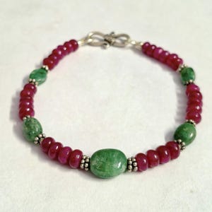 May include: A beaded bracelet featuring alternating sections of small, round, deep red beads and larger, smooth, oval-shaped green beads. The beads are separated by small, textured silver spacers, and the bracelet is secured with a silver S-hook clasp.