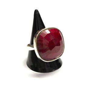 May include: A silver ring with a large, square-cut ruby gemstone. The ruby is a deep red colour and set in a silver band. The ring is displayed on a black cone-shaped stand against a white background.