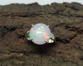 Puede incluir: Un anillo de plata con una gran piedra de ópalo redonda. El ópalo muestra una base blanca lechosa con destellos iridiscentes de color rosa, verde y amarillo. El engaste del anillo tiene cuatro garras.