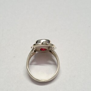 Puede incluir: Un anillo de plata con una piedra preciosa roja brillante y redonda. El anillo tiene una banda simple y un engaste decorativo alrededor de la piedra. La piedra preciosa tiene aproximadamente 1,3 cm de di&aacute;metro. El anillo est&aacute; sobre un fondo blanco liso.