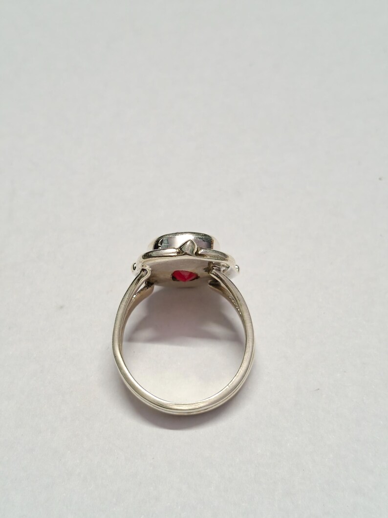 Puede incluir: Un anillo de plata con una piedra preciosa roja brillante y redonda. El anillo tiene una banda simple y un engaste decorativo alrededor de la piedra. La piedra preciosa tiene aproximadamente 1,3 cm de di&aacute;metro. El anillo est&aacute; sobre un fondo blanco liso.