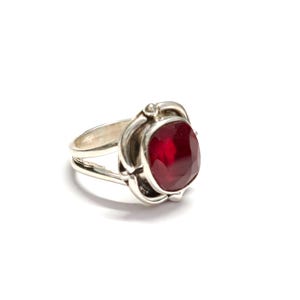 Puede incluir: Un anillo de plata con una gran gema roja oscura, facetada y de forma ovalada. La gema est&aacute; engastada en un engaste decorativo de inspiraci&oacute;n floral. El anillo tiene un dise&ntilde;o de doble banda y est&aacute; sobre un fondo blanco.