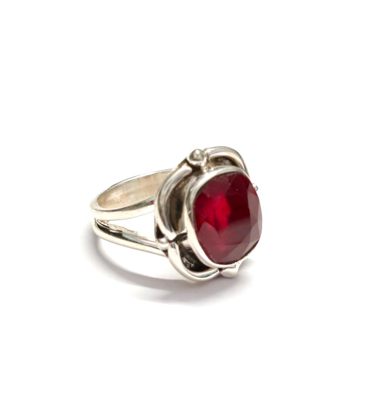 Puede incluir: Un anillo de plata con una gran gema roja oscura, facetada y de forma ovalada. La gema est&aacute; engastada en un engaste decorativo de inspiraci&oacute;n floral. El anillo tiene un dise&ntilde;o de doble banda y est&aacute; sobre un fondo blanco.