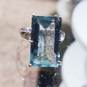 May include: A silver ring with a large, rectangular, light blue gemstone. The gemstone has a faceted cut and is set in a prong setting.