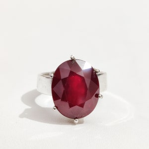 May include: A silver ring with a large, oval-cut, deep red gemstone. The faceted gemstone is the focal point, reflecting light. The ring has a simple, polished band and small prongs securing the stone. The ring is a statement piece.