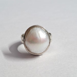 Peut inclure: Bague en argent ornée d'une grosse perle blanche nacrée. La perle, de forme irrégulière, est sertie dans un sertissage en argent. La monture de la bague est également en argent, pour un design classique et élégant. La perle mesure environ 2,5 cm.