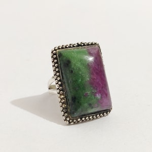 Op de afbeelding: Een zilveren ring met een rechthoekige groene en roze edelsteen, gezet in een parelrand.