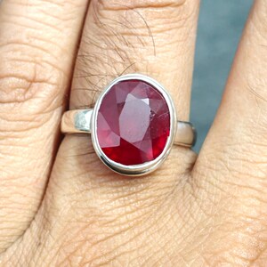 May include: A silver ring with a large, oval-cut ruby gemstone. The ruby is a deep red colour and is set in a simple, polished silver band. The ring is shown on a finger.