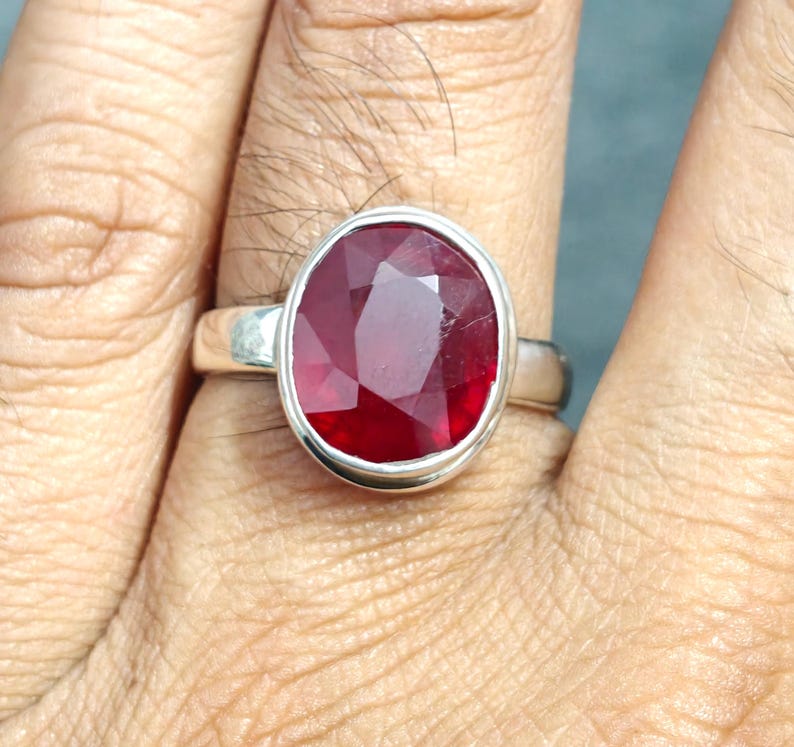 May include: A silver ring with a large, oval-cut ruby gemstone. The ruby is a deep red colour and is set in a simple, polished silver band. The ring is shown on a finger.