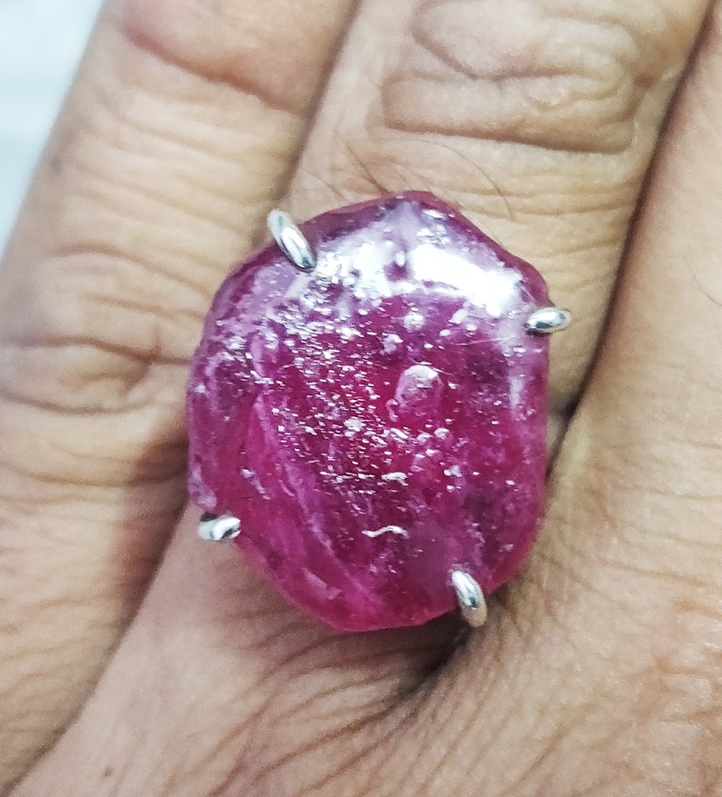 Pode incluir: Um anel com uma grande gema de rubi oval rosa engastada em uma banda de prata. O rubi tem um aspecto &aacute;spero e natural e &eacute; rodeado por quatro garras de prata.