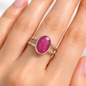 Op de afbeelding: Een zilveren ring met een grote, ovaal geslepen robijn. De ring heeft een dubbele band, met de robijn in een bezel gezet. De robijn heeft een levendige roze-rode tint. De ring wordt aan een vinger getoond.