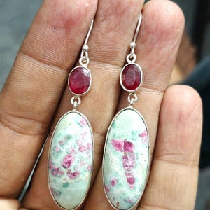 Pu&ograve; includere: Un paio di orecchini in argento. Ogni orecchino ha una piccola gemma rossa ovale sopra una gemma pi&ugrave; grande, ovale, verde chiaro con inclusioni rosa. Gli orecchini pendono da ganci d'argento.