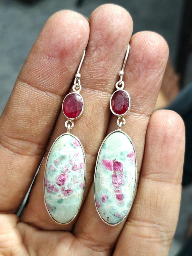 Pu&ograve; includere: Un paio di orecchini in argento. Ogni orecchino ha una piccola gemma rossa ovale sopra una gemma pi&ugrave; grande, ovale, verde chiaro con inclusioni rosa. Gli orecchini pendono da ganci d'argento.