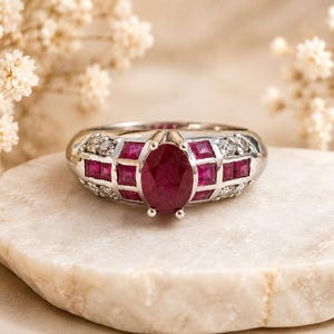 May include: A silver-toned ring featuring a central oval-cut deep red ruby, flanked by a row of square-cut rubies and small round clear stones set into the band. The metal band has a polished finish and a wide, structured design.