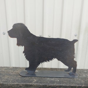 May include: A black metal silhouette of a Cocker Spaniel dog standing on a black metal base. The silhouette is cut out of metal and has a rustic finish.