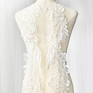 Op de afbeelding: Witte kant applique met bloemen- en bladmotieven. De kant is delicaat en ingewikkeld en zou perfect zijn om een ​​toets van elegantie toe te voegen aan een bruidsjurk of andere kleding voor speciale gelegenheden.