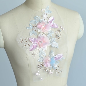 May include: A delicate floral applique with pink, blue, and white flowers and leaves. The applique is adorned with sparkling rhinestones and pearls, making it perfect for adding a touch of elegance to clothing or crafts.