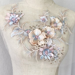 May include: A floral applique featuring embroidered flowers in peach, pink, and blue, embellished with tiny beads. The applique is displayed on a mannequin, highlighting its design and texture. The delicate details and colour palette suggest a craft or sewing project.