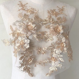 May include: A delicate, floral lace applique in shades of gold and cream. The design features embroidered flowers, leaves, and pearl-like beads. The applique is likely for embellishing garments or crafts.