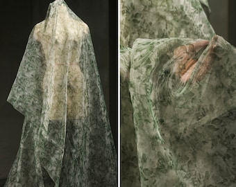 May include: Green floral patterned sheer fabric. The fabric is draped over a dress form and held in a hand.