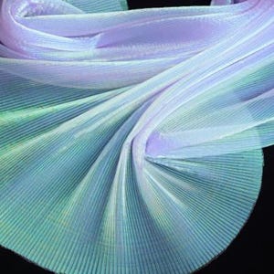 Könnte beinhalten: Ein fließender, plissierter Stoff in Blau-, Grün- und Lilatönen. Der Stoff ist in weichen Falten angeordnet, wodurch ein Gefühl von Bewegung und Textur entsteht. Das schillernde Material fängt das Licht ein und verleiht ihm ein schimmerndes Aussehen.