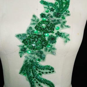 May include: An emerald green floral applique patch featuring intricate beadwork, sequins, and embroidery on a sheer mesh base. The design showcases layered flower motifs and leafy vine patterns, creating a textured, decorative embellishment.