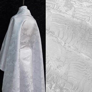 May include: White fabric with a detailed, wave-like pattern. The fabric is draped over a form, showcasing its texture and design. The close-up reveals the intricate details of the woven pattern, suitable for apparel or home decor.