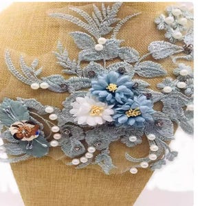 May include: An intricate floral applique with embroidered leaves and flowers in blue, white, and green hues. The design is adorned with tiny pearls and rhinestones, offering a textured and elegant appearance. This applique is suitable for various craft projects.