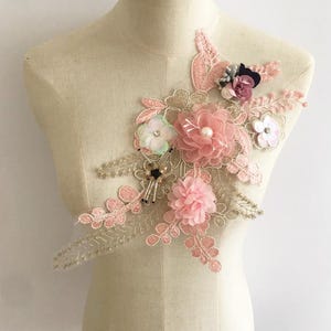 May include: Pink and gold floral applique with pearl accents. This delicate lace embellishment features a variety of flower shapes and textures, perfect for adding a touch of elegance to clothing or crafts.